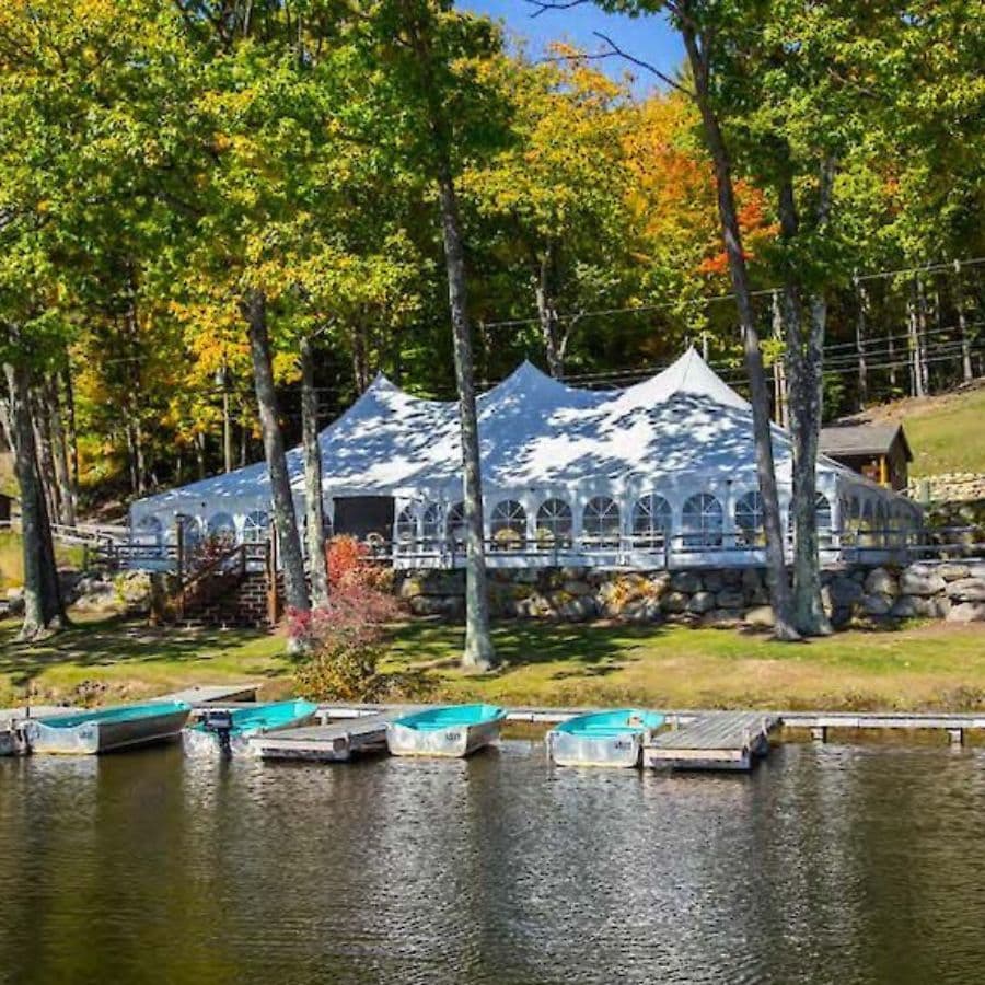 A large white tent sits near a lake, surrounded by colorful trees and boats on the water.