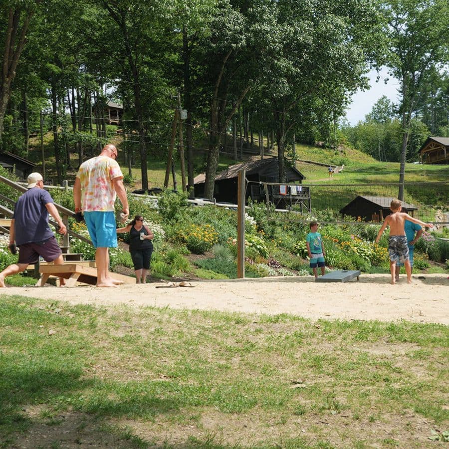A group of people engages in outdoor activities on a sandy area surrounded by greenery and wooden structures.