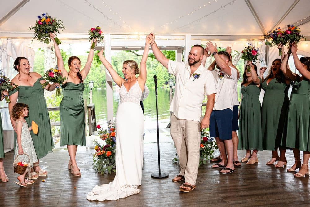 A bride and groom celebrate with their wedding party, raising their arms in joy under a decorated tent.