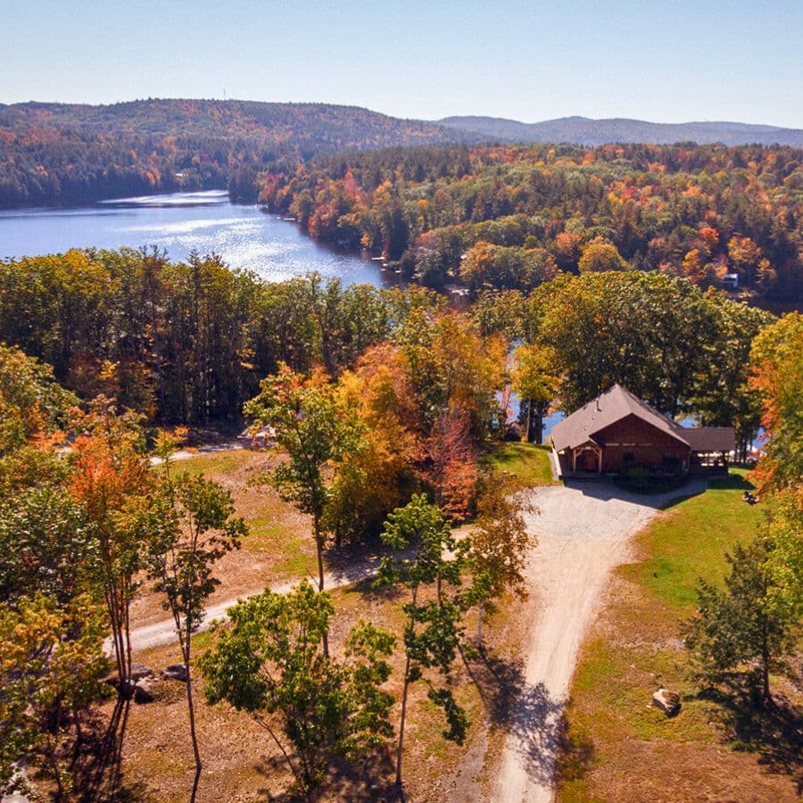 Aerial view of a lake surrounded by autumn-colored trees and a cabin beside a gravel path.