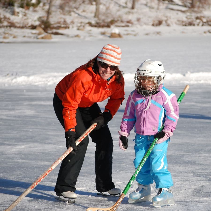A woman and a young girl smile while ice skating on a frozen surface, both holding hockey sticks.