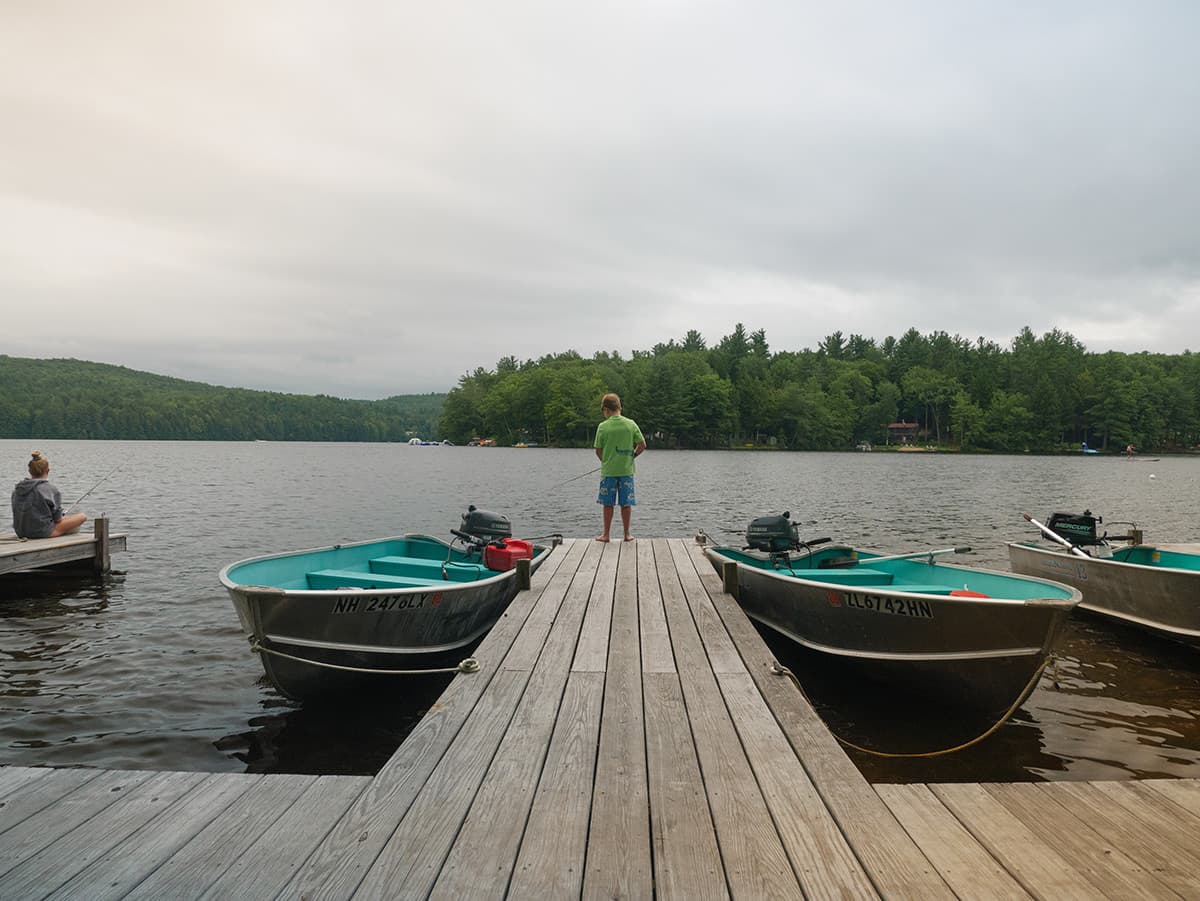 A young person stands on a wooden dock by a lake, facing the water, while another person sits and fishes nearby.