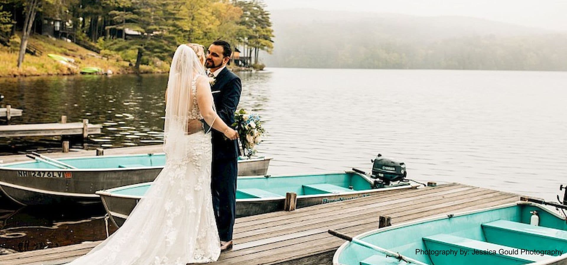 A bride and groom embrace on a dock by a tranquil lake, surrounded by boats.