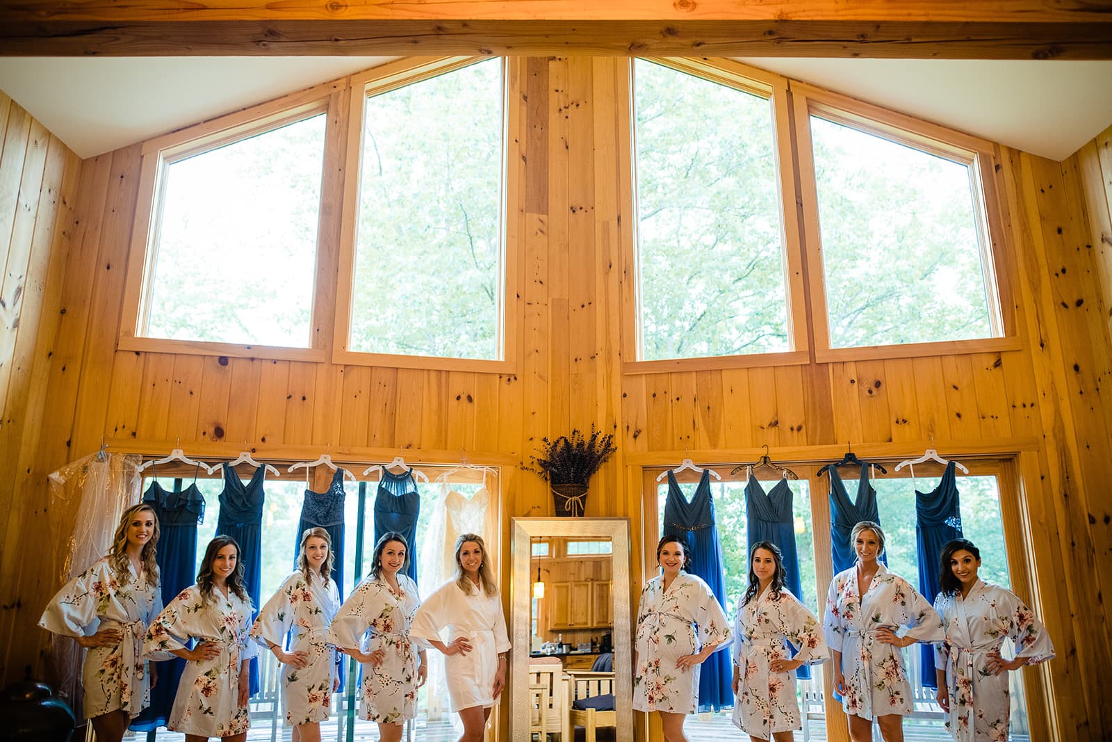 Bridesmaids in floral robes pose in a wooden cabin with blue dresses hanging in the background.
