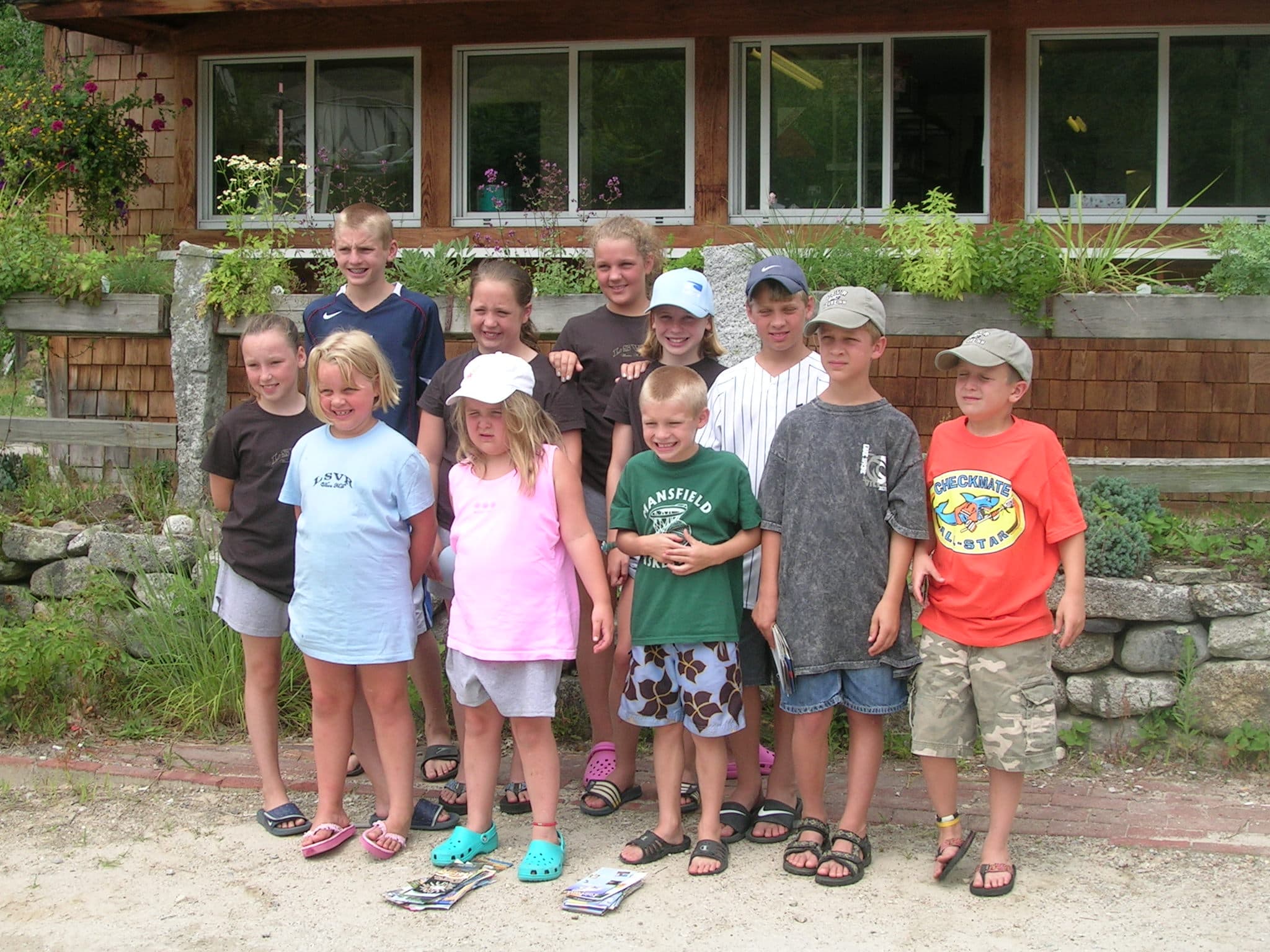 A group of ten children posing together outdoors in front of a wooden building.