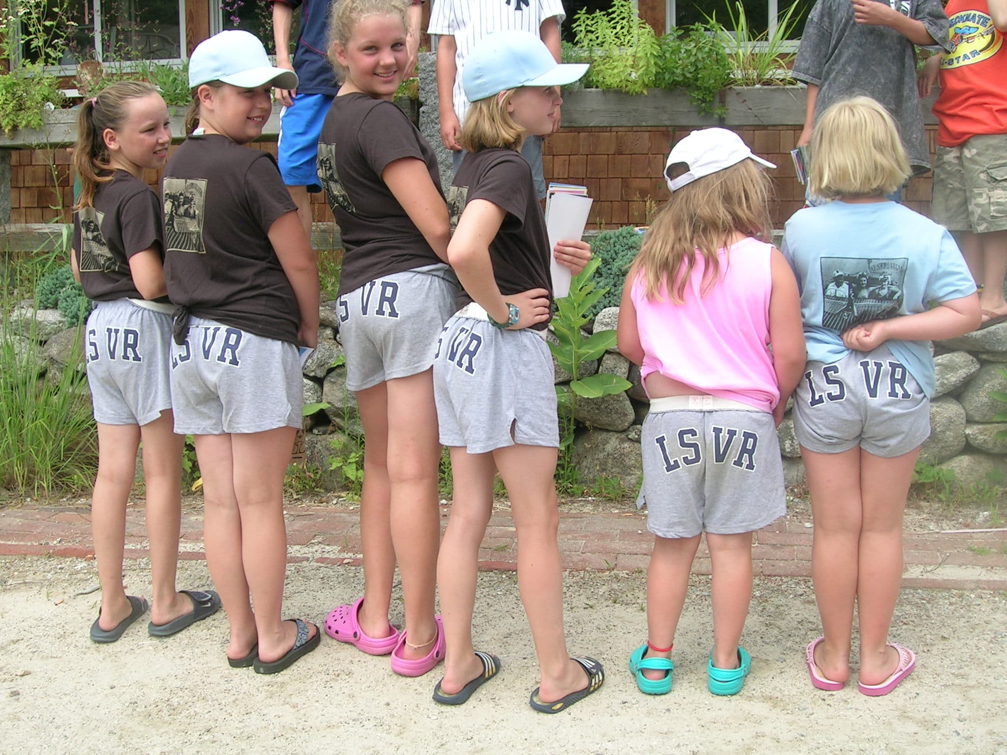 A group of girls in matching grey shorts and T-shirts stand together, showcasing the back of their outfits.