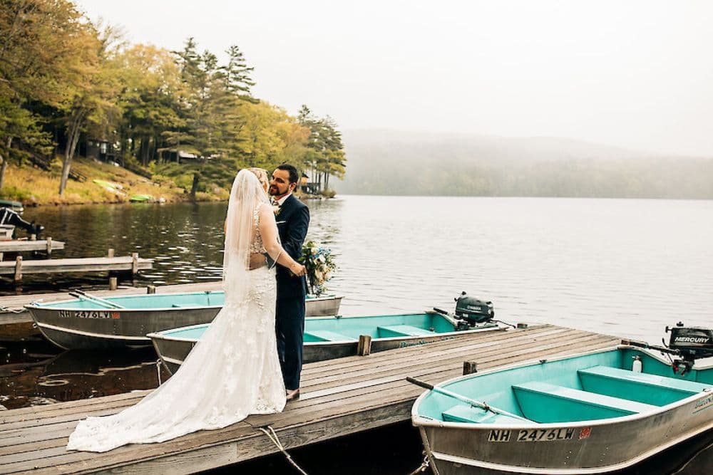 A couple in wedding attire shares a romantic moment on a dock by a lake, surrounded by boats and misty mountains.