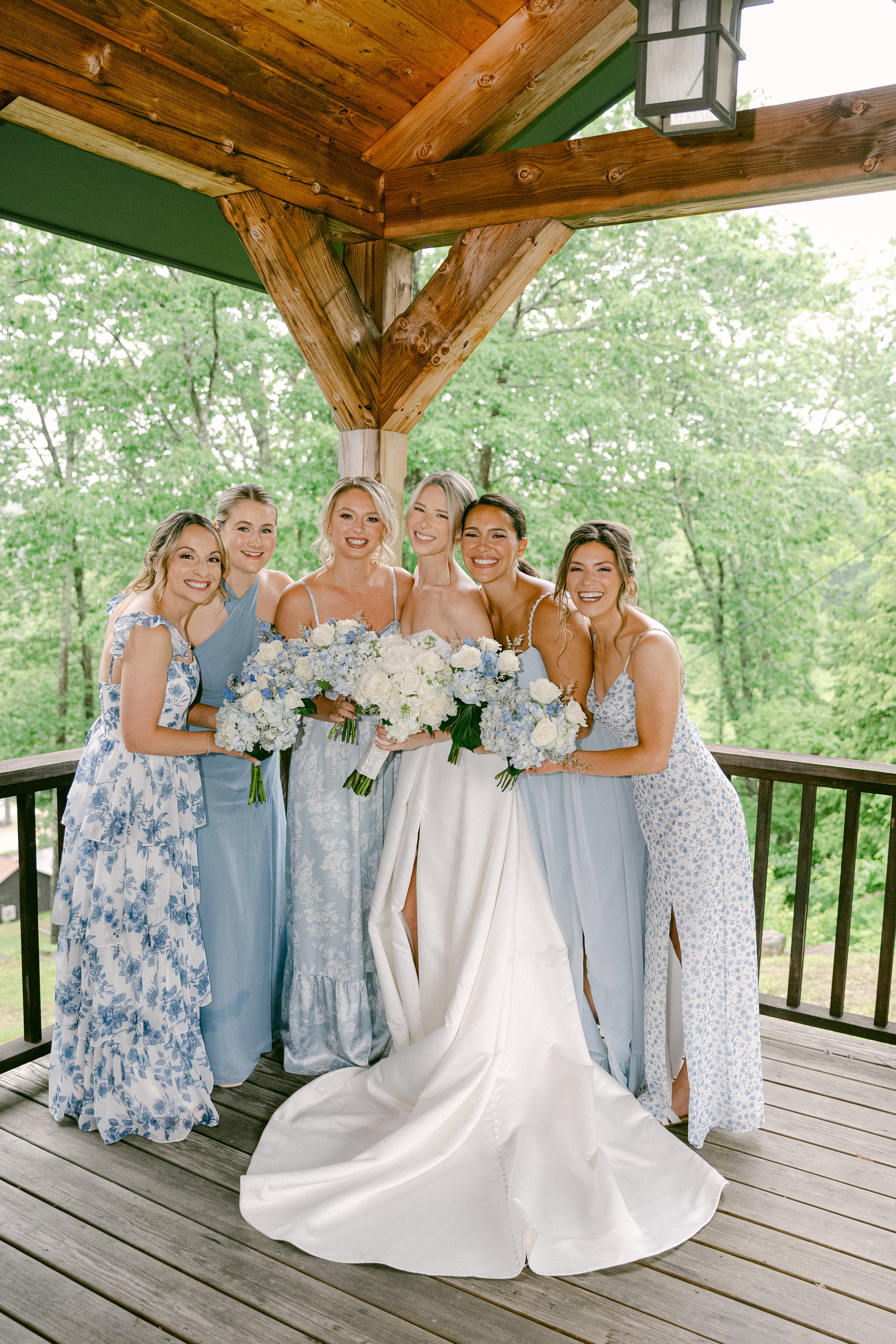 A group of six women in blue dresses smile together on a porch, holding bouquets of flowers.