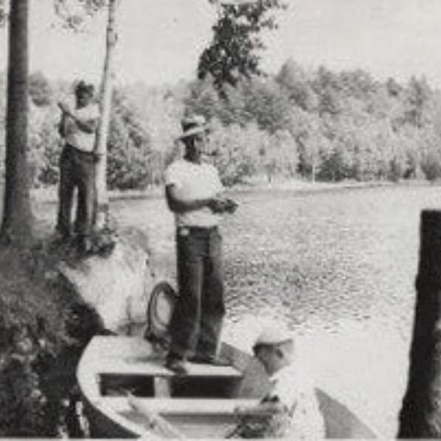 Three people fishing by a lake, one on a boat and two standing nearby.
