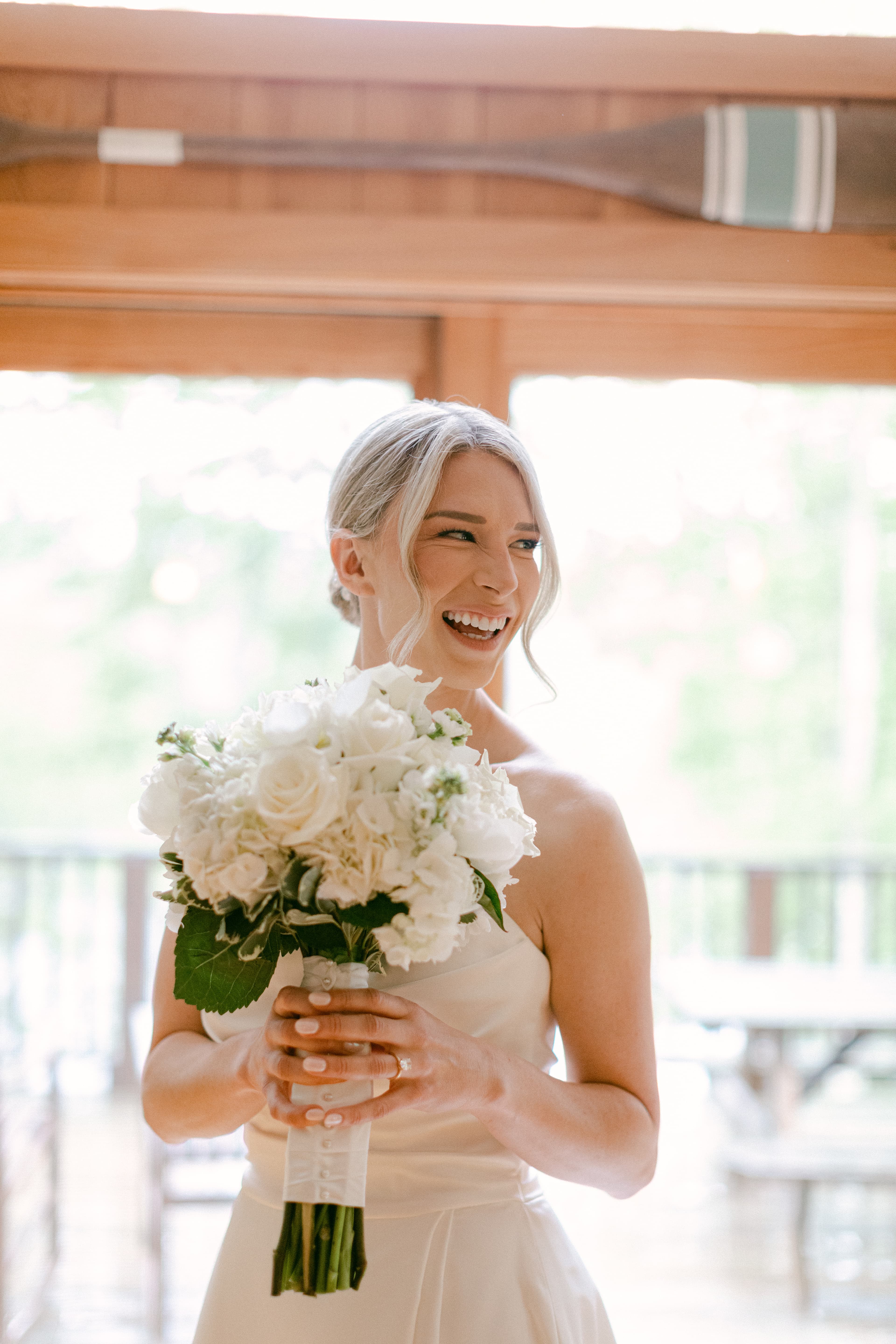 A smiling bride in a white dress holds a bouquet of white flowers indoors.