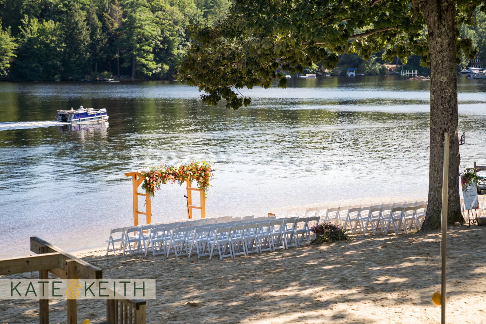 A lakeside wedding setup featuring a floral arch and rows of white chairs on the sandy beach.