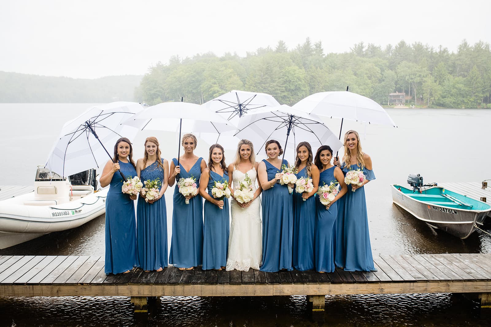 Bridesmaids in blue dresses holding bouquets and umbrellas pose on a dock by a lake.