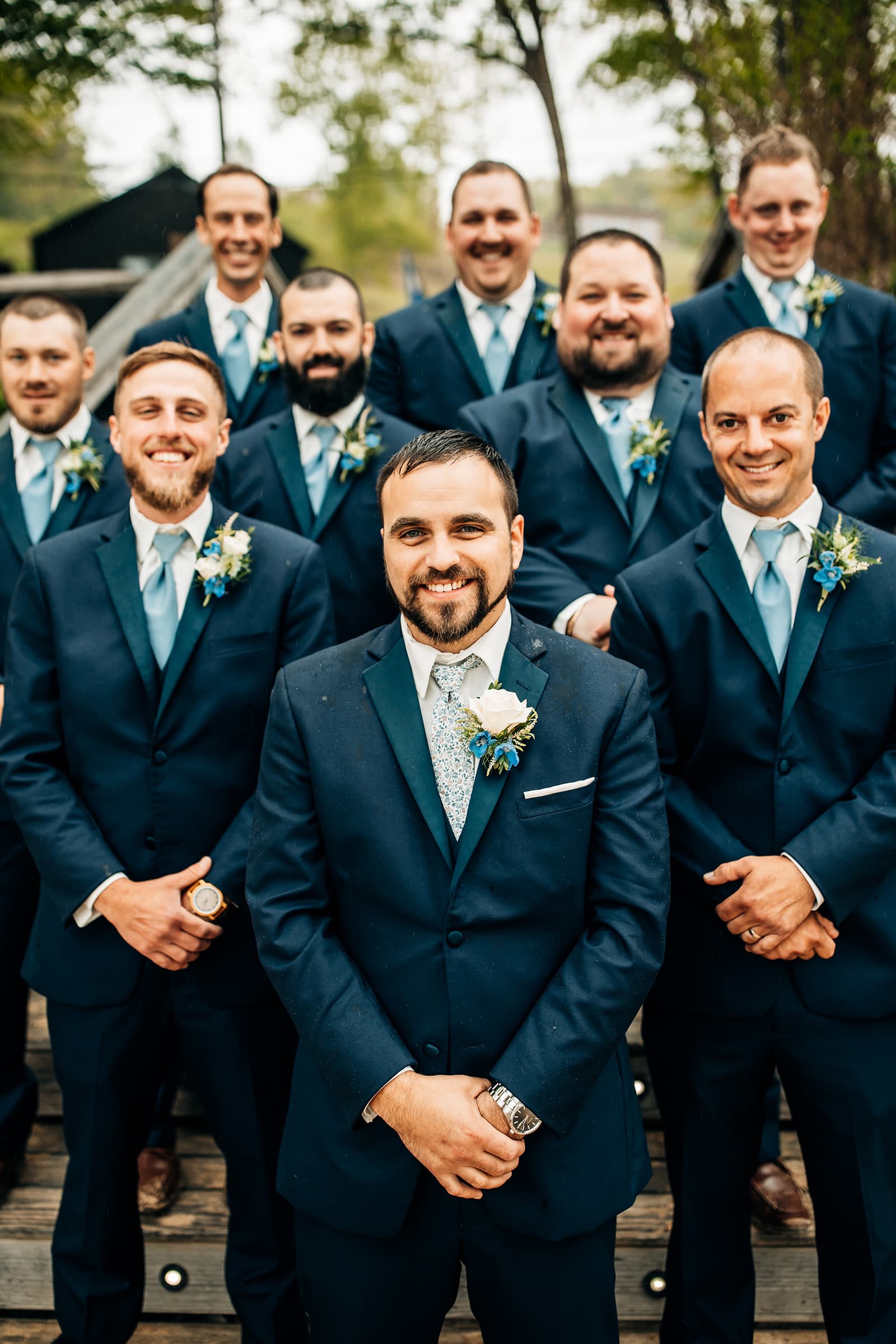 A group of groomsmen in navy suits and light blue ties pose together outdoors.