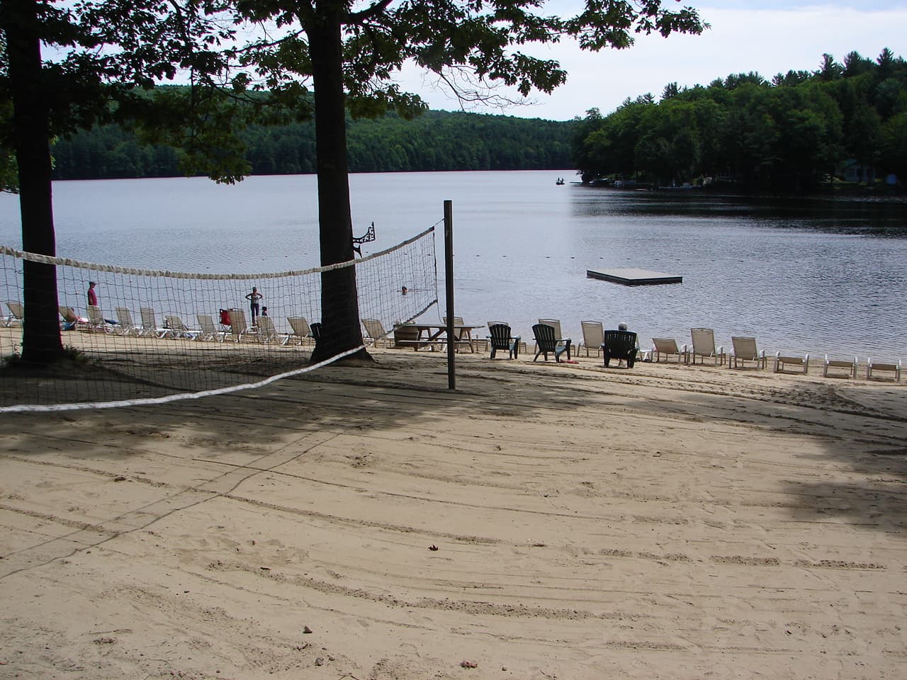 A sandy beach with a volleyball net, lounge chairs, and a calm lake surrounded by trees.