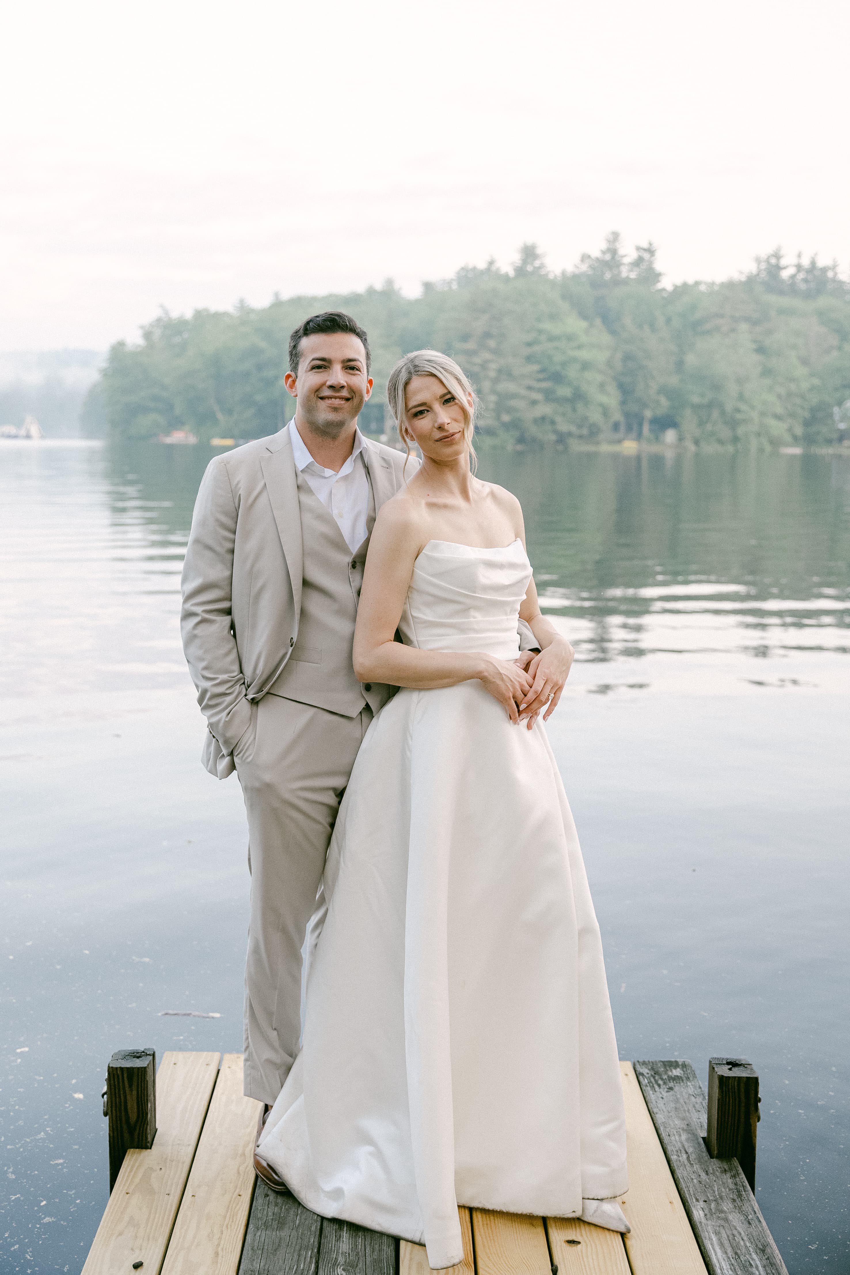 A bride and groom pose together on a dock by a lake, surrounded by lush greenery and calm waters.