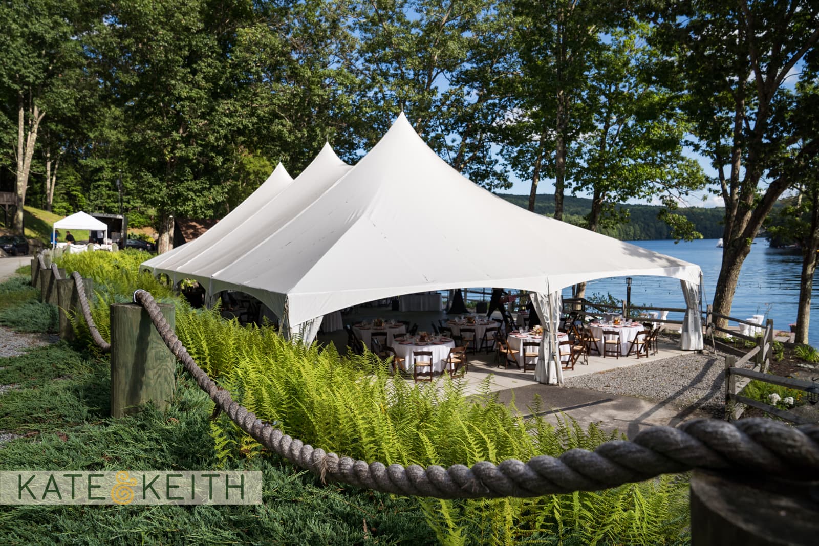 A white tent with tables set up is situated near a lake, surrounded by greenery and trees.