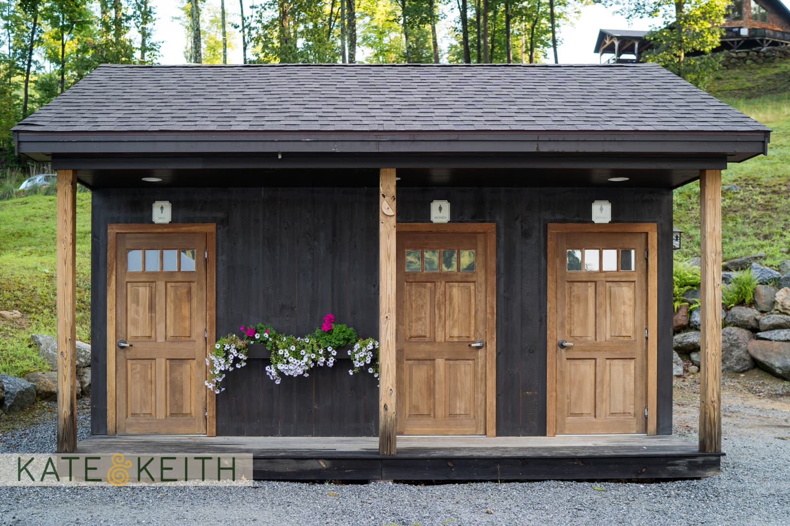 Two wooden doors on a black building façade with flower boxes beneath, surrounded by trees.