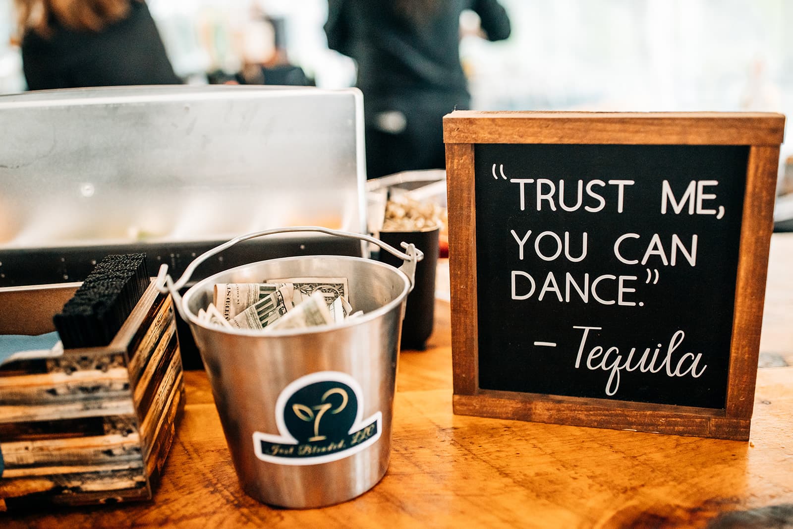 A sign reads, "Trust me, you can dance," attributed to Tequila, next to a bucket of cash and a brush holder on a wooden table.