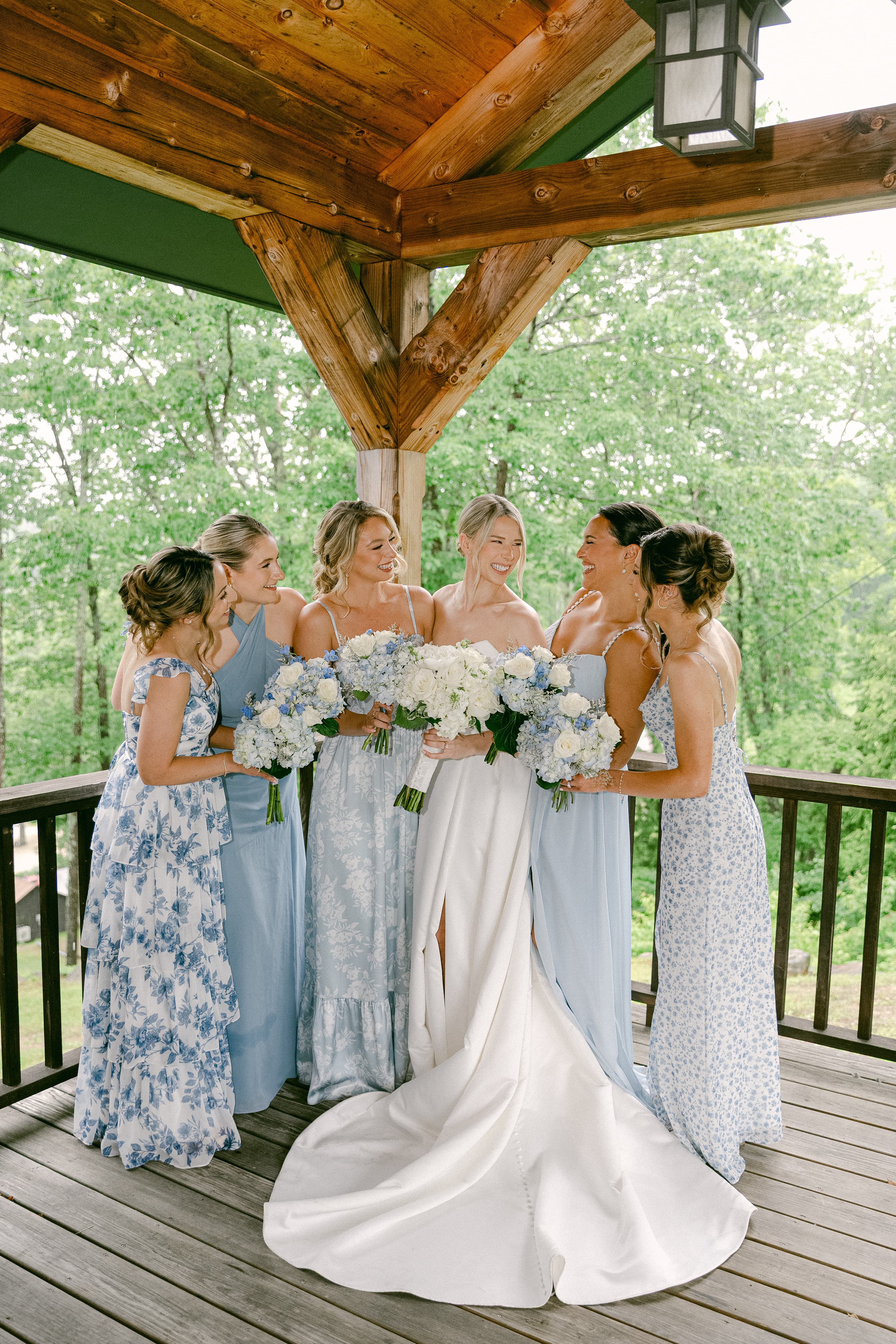 A joyful group of bridesmaids in light blue dresses gather around the bride, who wears a white gown, all holding white and blue bouquets under a covered wooden pavilion.