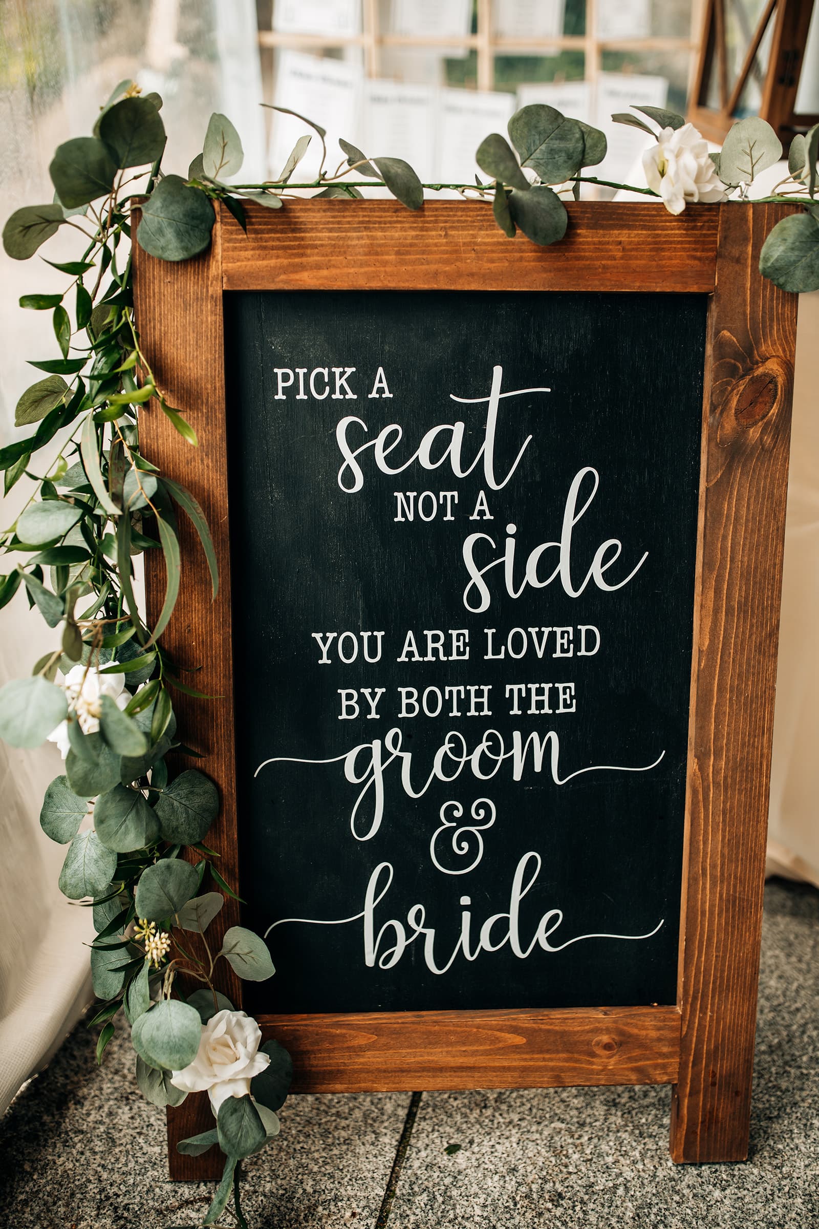 A wooden sign with greenery that reads, "Pick a seat, not a side; you are loved by both the groom & bride."