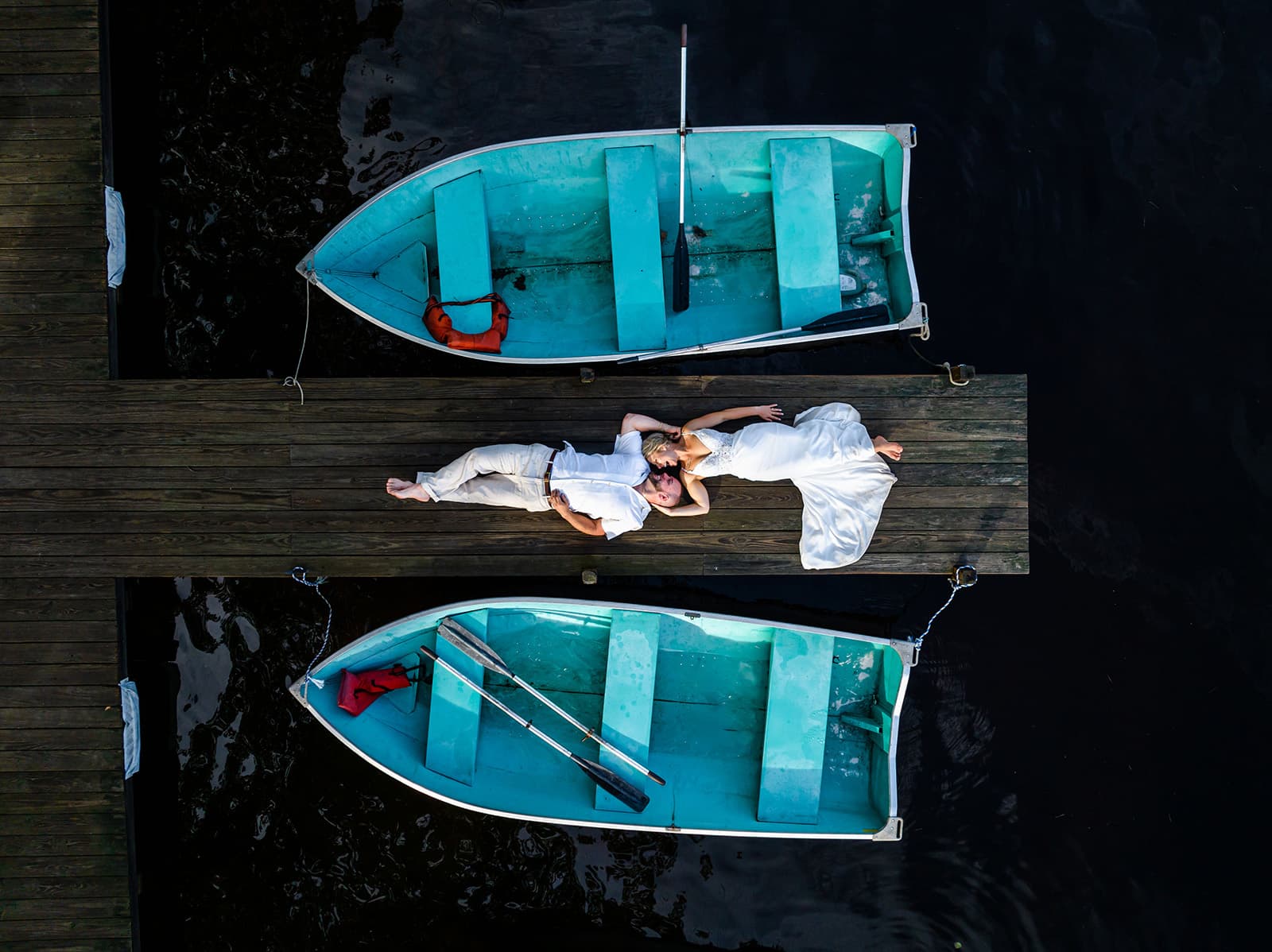 A couple lies on a wooden dock between two blue boats, surrounded by dark water.