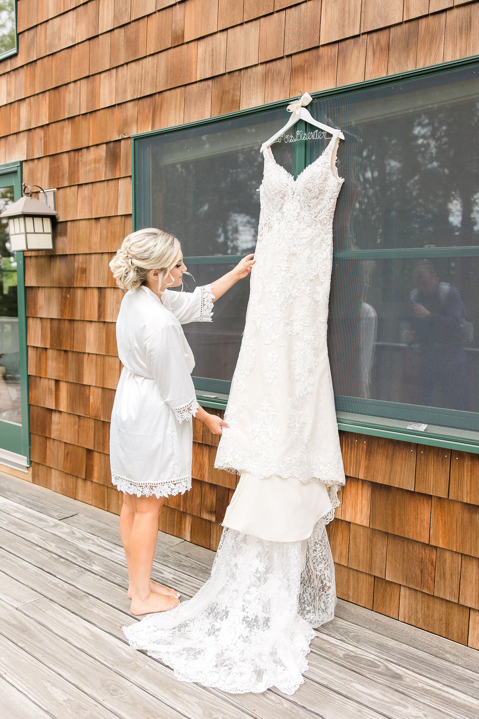 A bride in a white robe admires her wedding dress hanging against a wooden wall.
