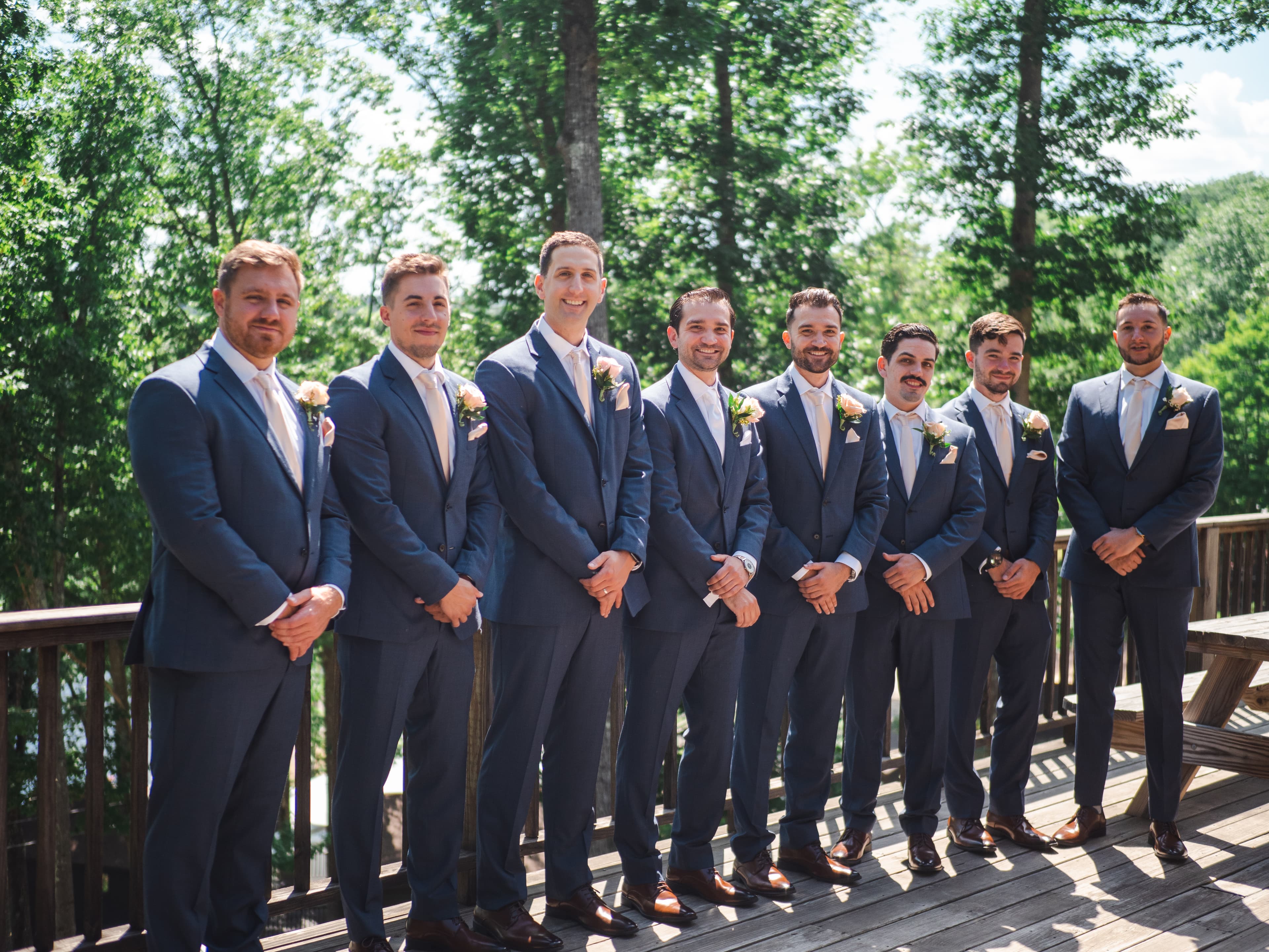 A group of eight men in matching navy suits stand on a deck surrounded by trees.