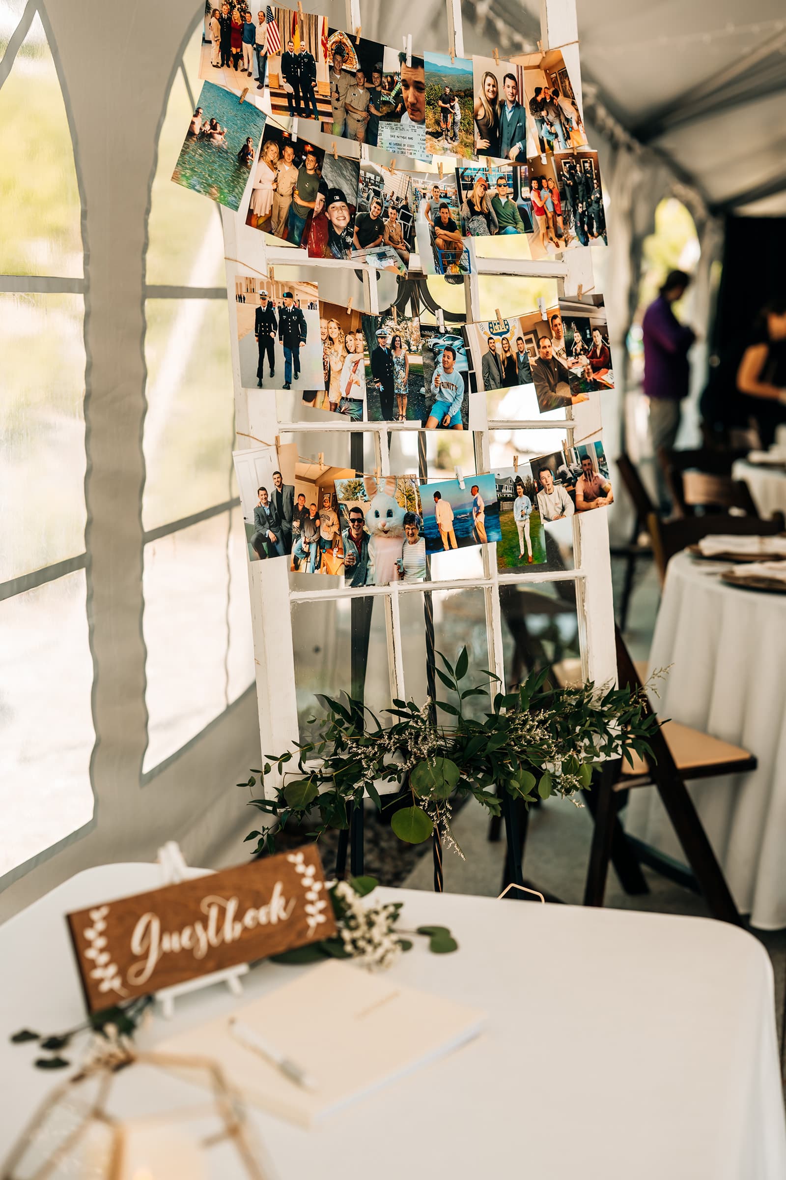 A display of photographs hangs on a wooden frame inside a tent, with a guestbook and greenery on a nearby table.