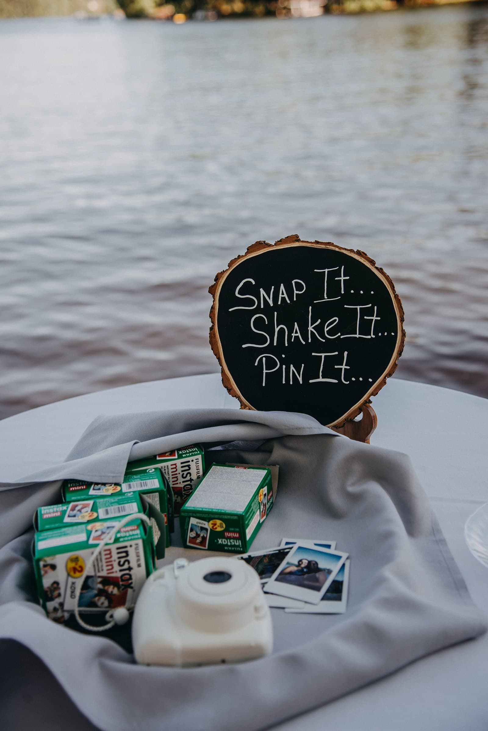 A table with a chalkboard sign and several boxes of instant film next to a camera, set against a lake backdrop.