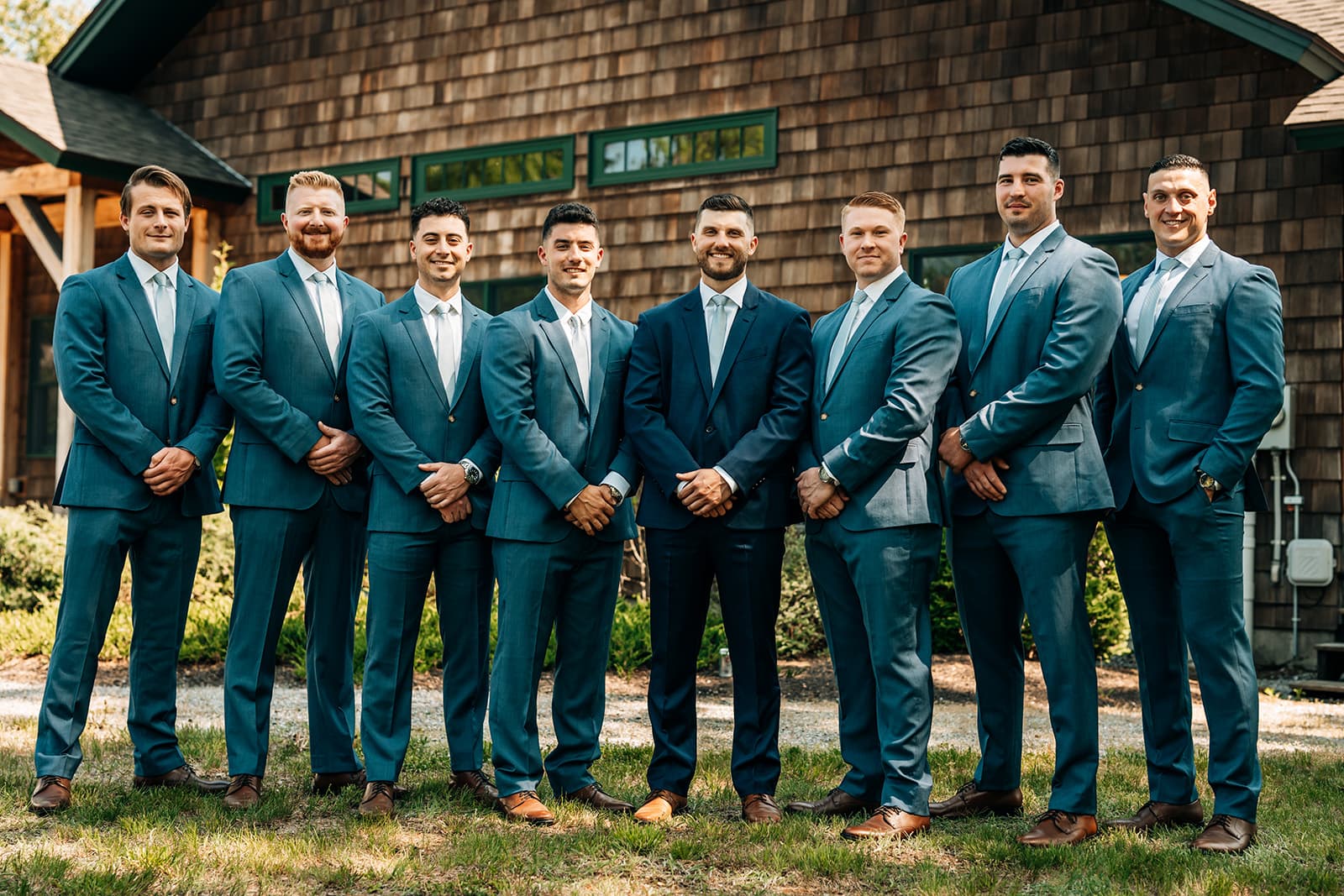 A group of eight men in matching blue suits pose together outside a wooden building.