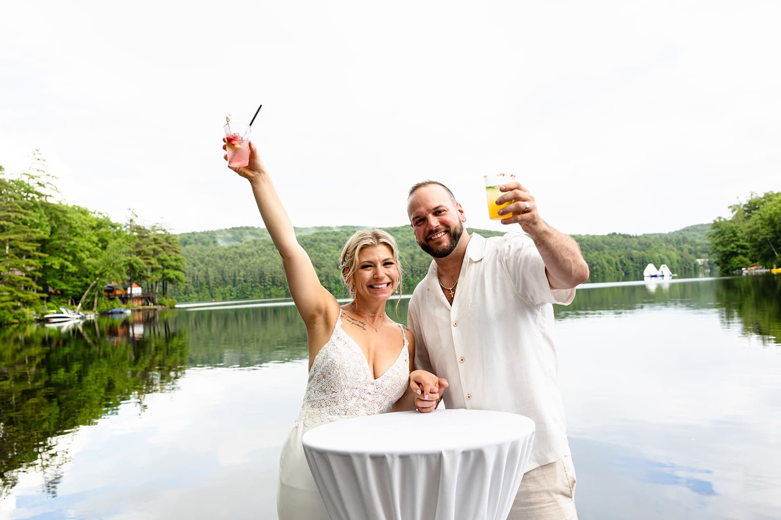 A smiling couple raises drinks in celebration by a serene lakeside.