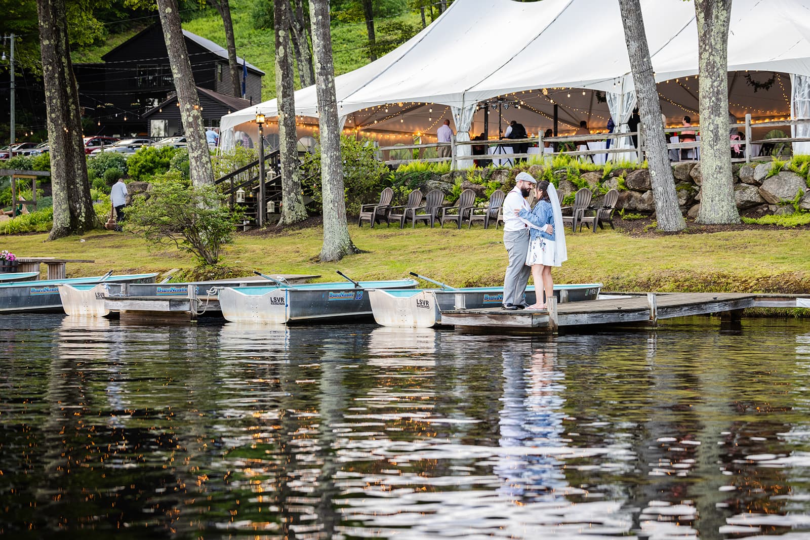 A couple shares a kiss on a dock by a tranquil lake, with a tented event space and boats nearby.