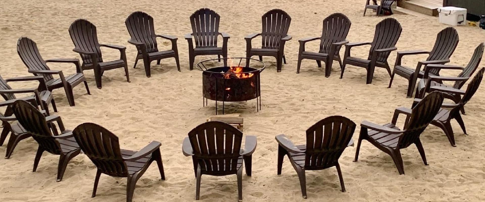 A circle of wooden chairs surrounds a fire pit on a sandy beach.