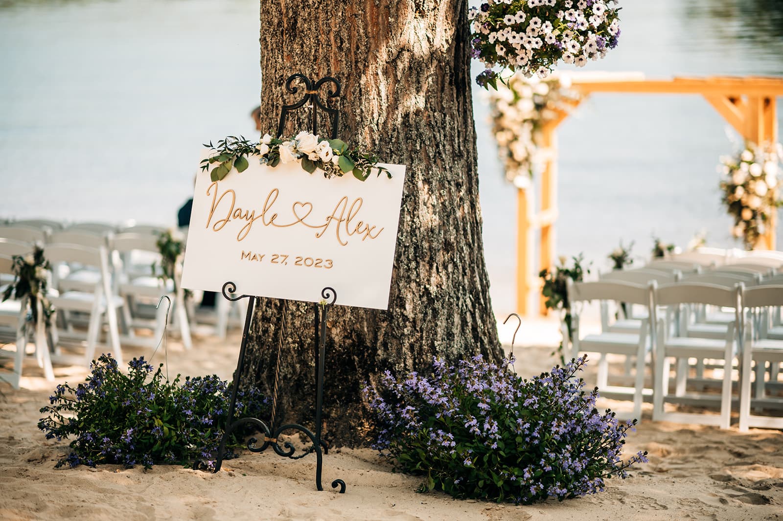 A wedding sign decorated with flowers announces "Dayle ♥ Alex, May 27, 2023" beside a tree near a sandy waterfront.