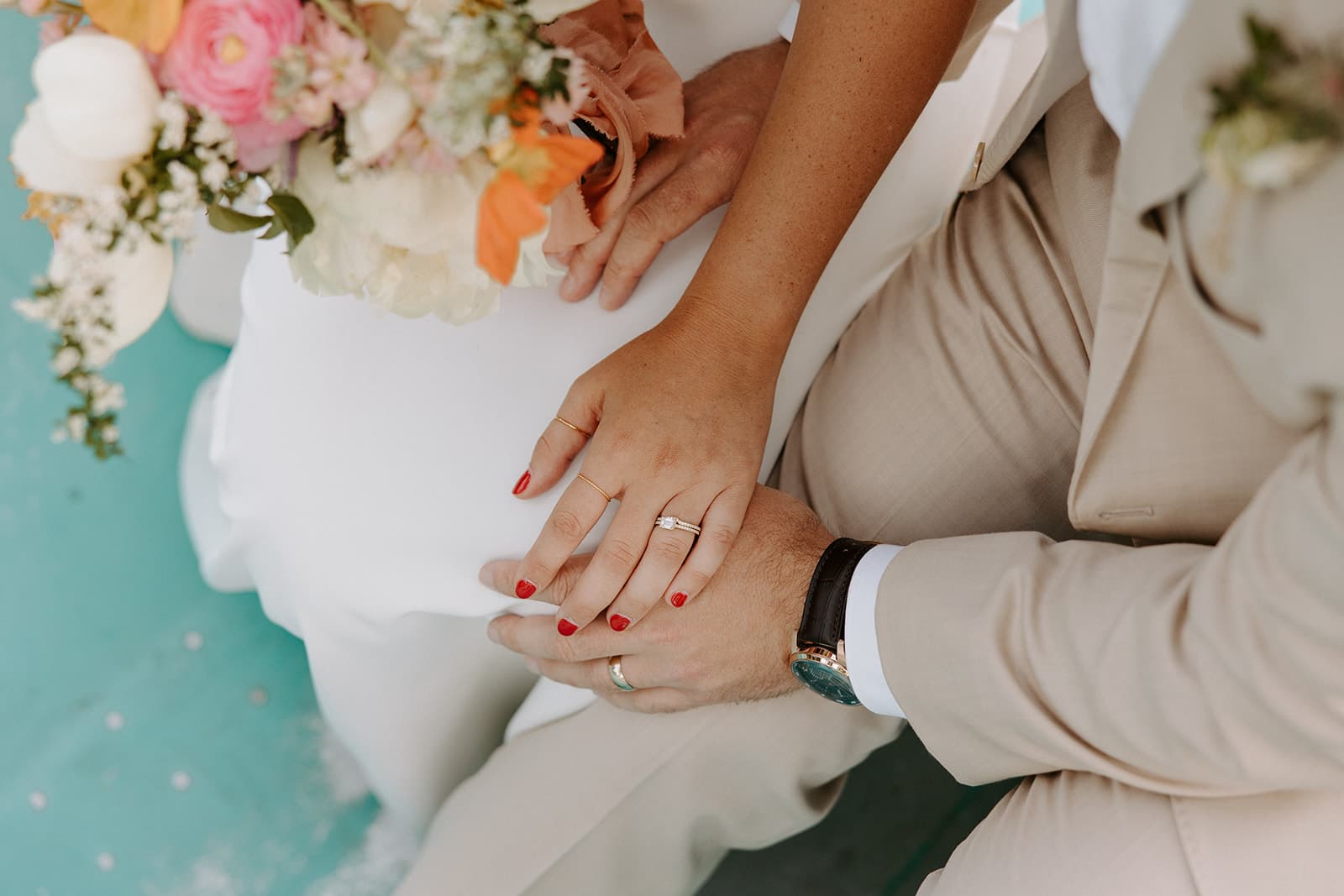 A close-up of two hands, one adorned with a ring and the other with red nail polish, resting together, with a bouquet in the background.