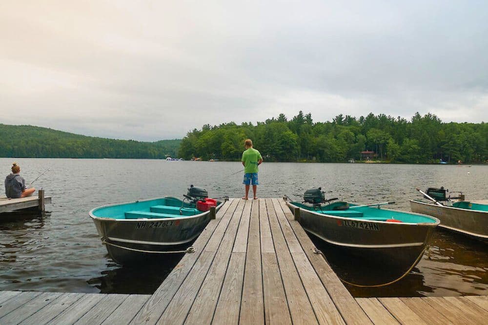 A boy stands on a wooden dock by a lake, while another person fishes nearby.