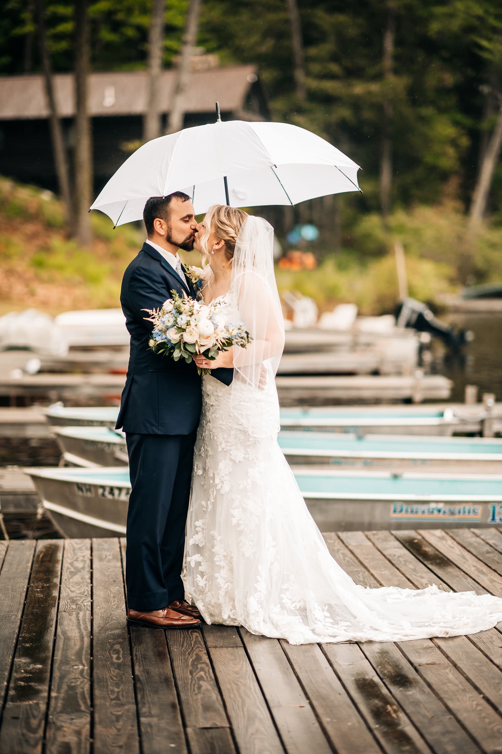 A bride and groom kiss under an umbrella on a wooden dock, surrounded by boats and nature.