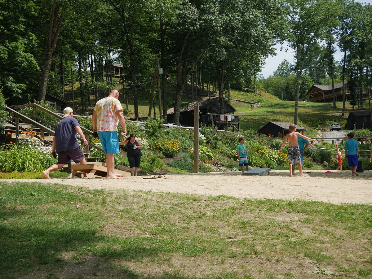 A group of children and adults play outdoors in a grassy area surrounded by trees and cabins.