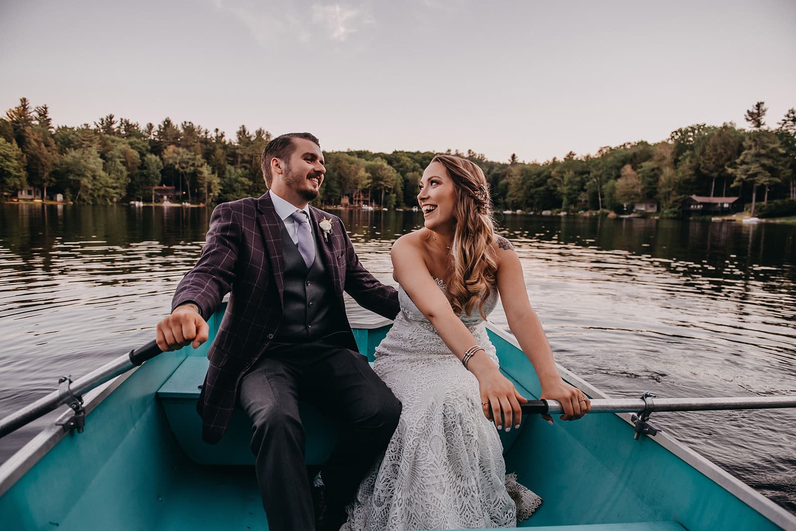 A smiling couple in formal attire rows a small boat on a calm lake surrounded by trees.