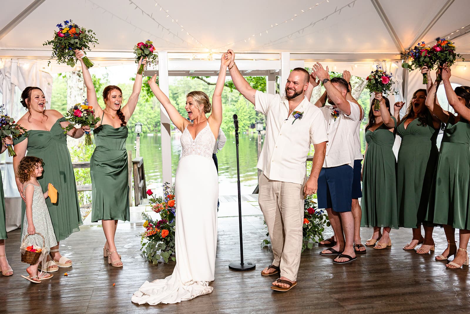 A joyful bride and groom celebrate with their wedding party under a tent, surrounded by flowers and greenery.