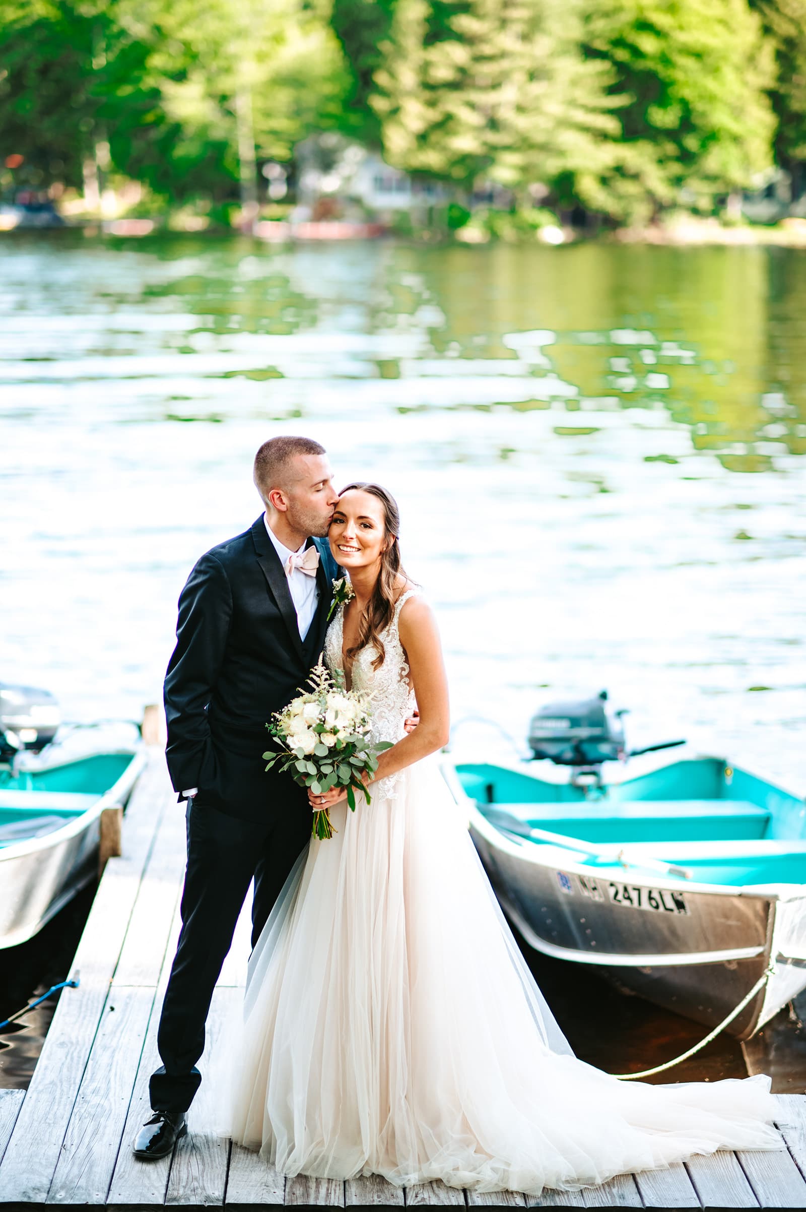 A bride and groom share a kiss on a dock by a lake, surrounded by boats and greenery.