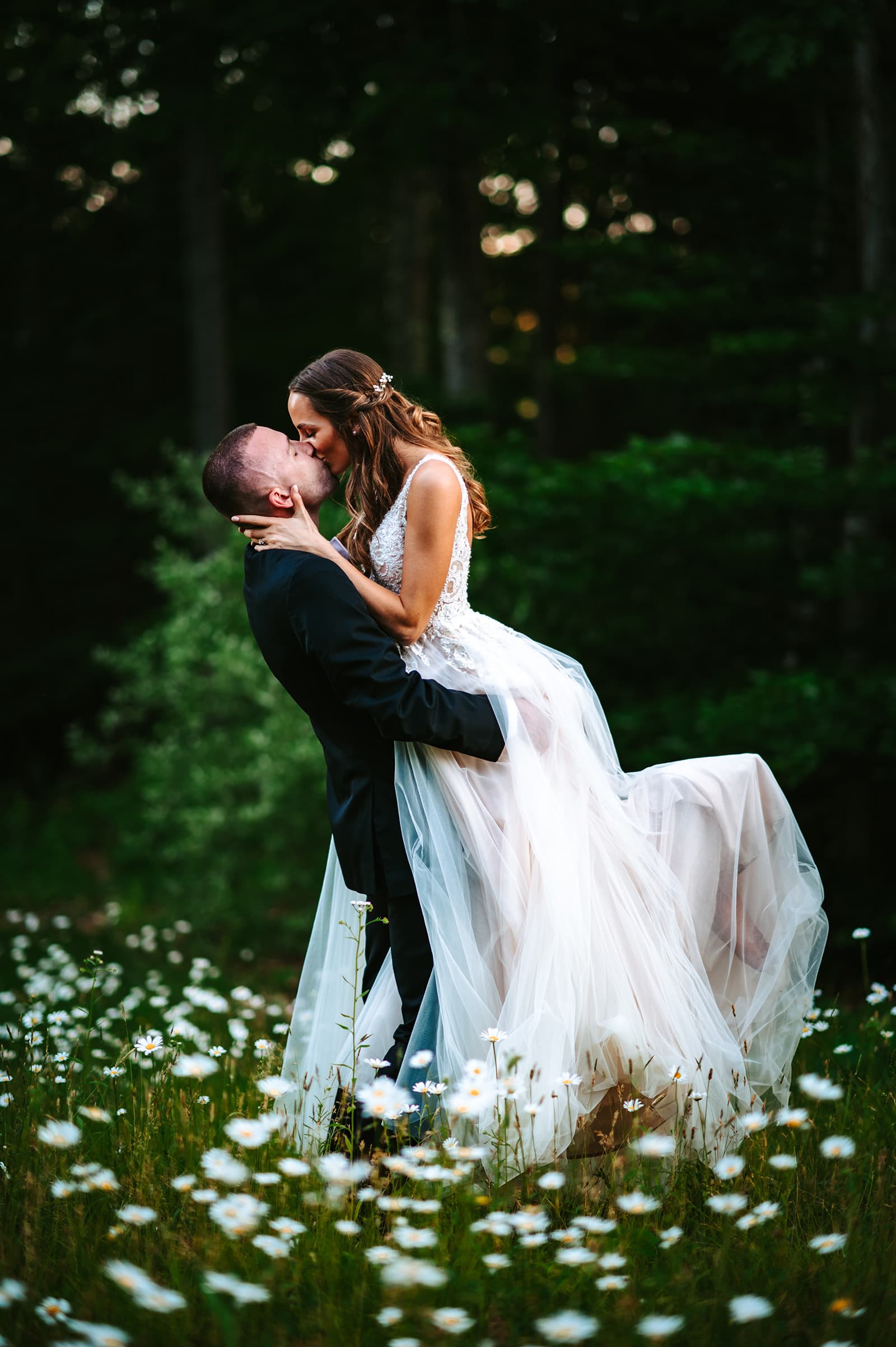 A couple shares a romantic kiss in a field of flowers, with the woman in a flowing wedding dress.