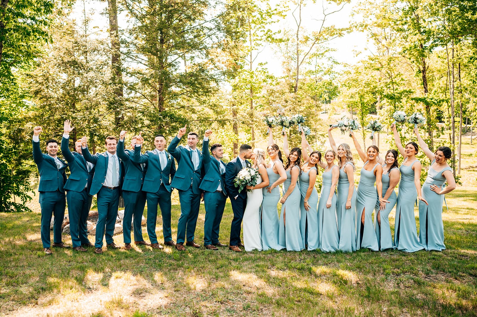 A wedding party joyfully poses outdoors, celebrating with raised fists and bouquets.