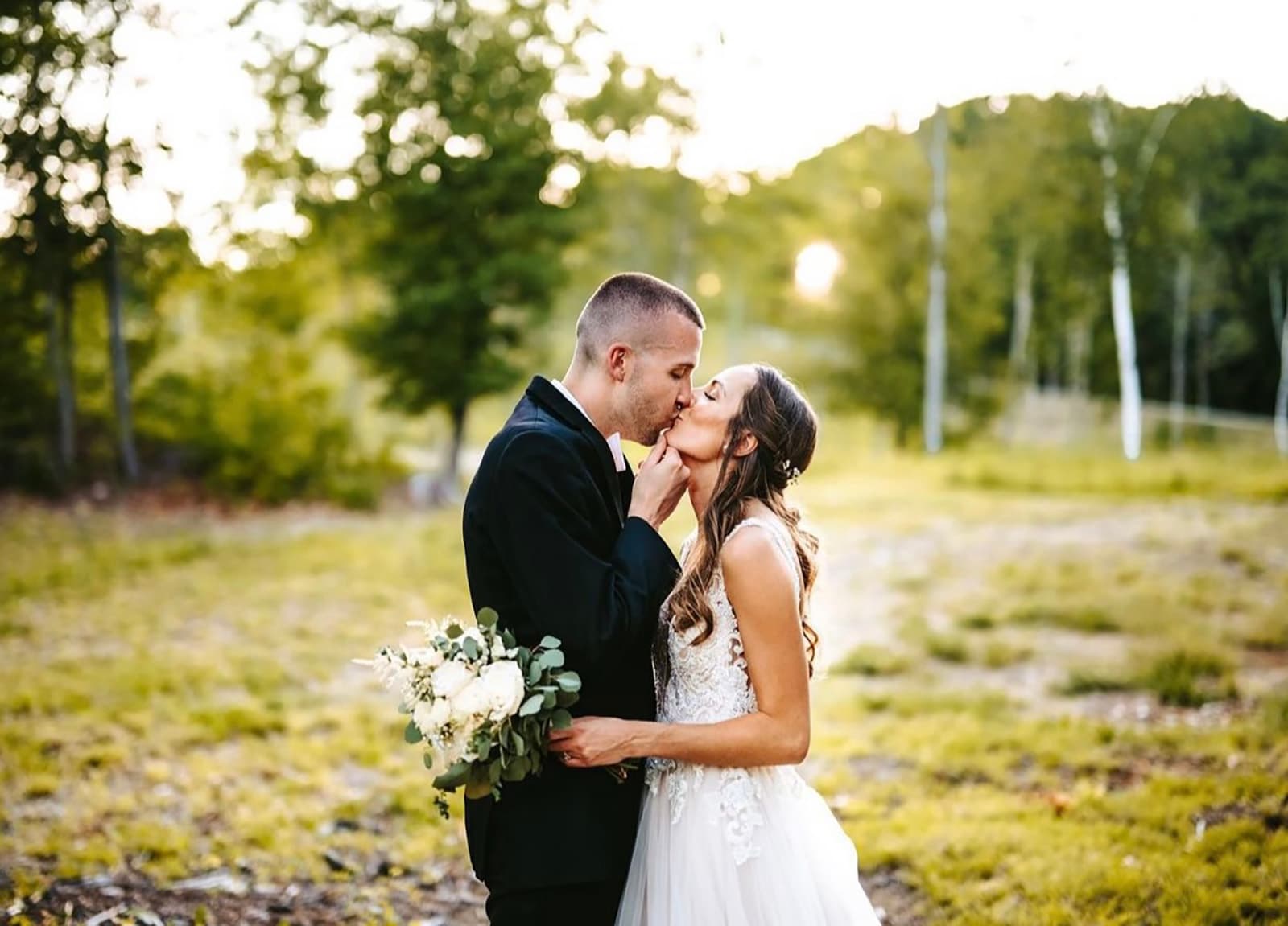 A bride and groom kiss while holding a bouquet in a sunny outdoor setting.