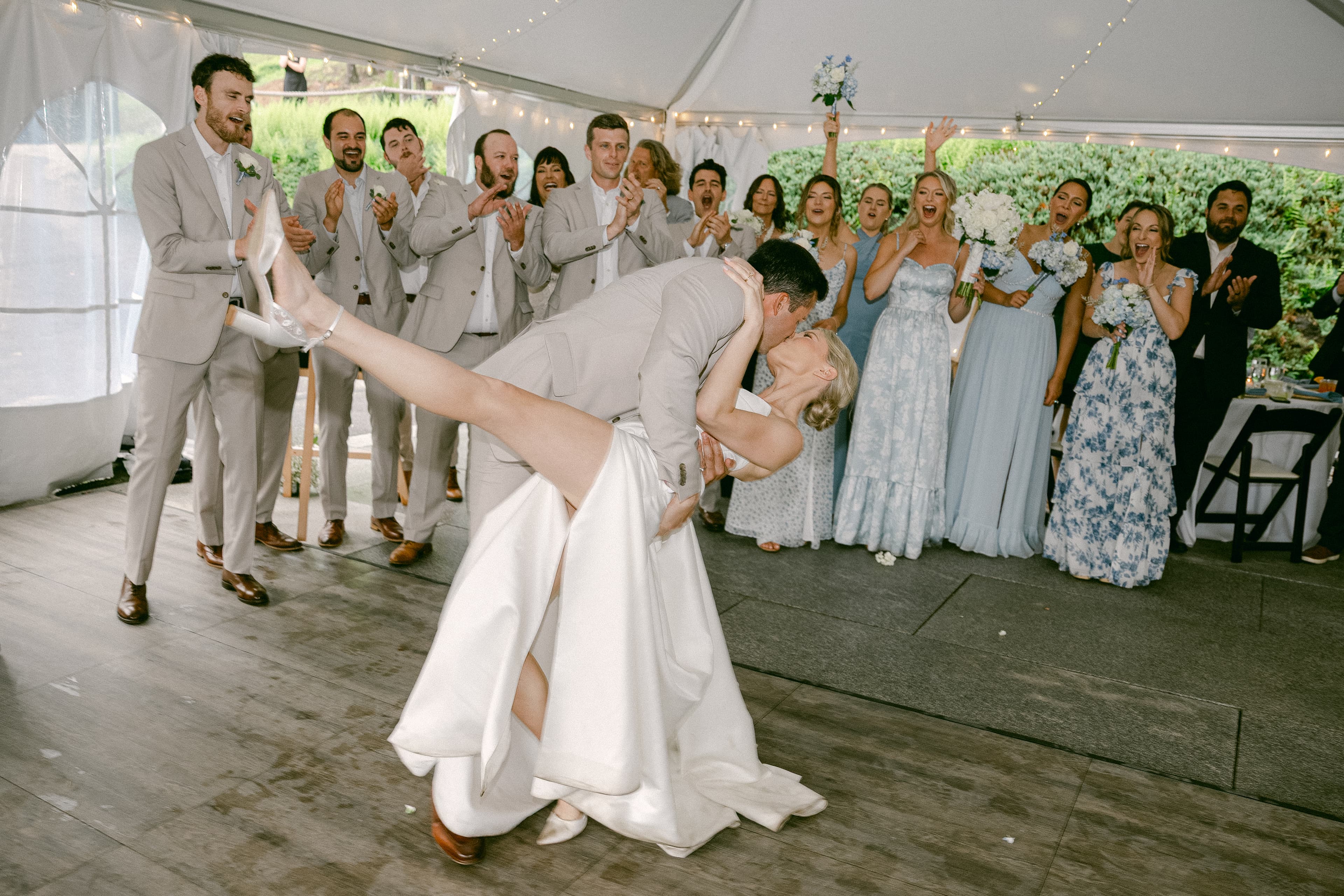 A bride and groom dance joyfully under a tent, surrounded by cheering guests.