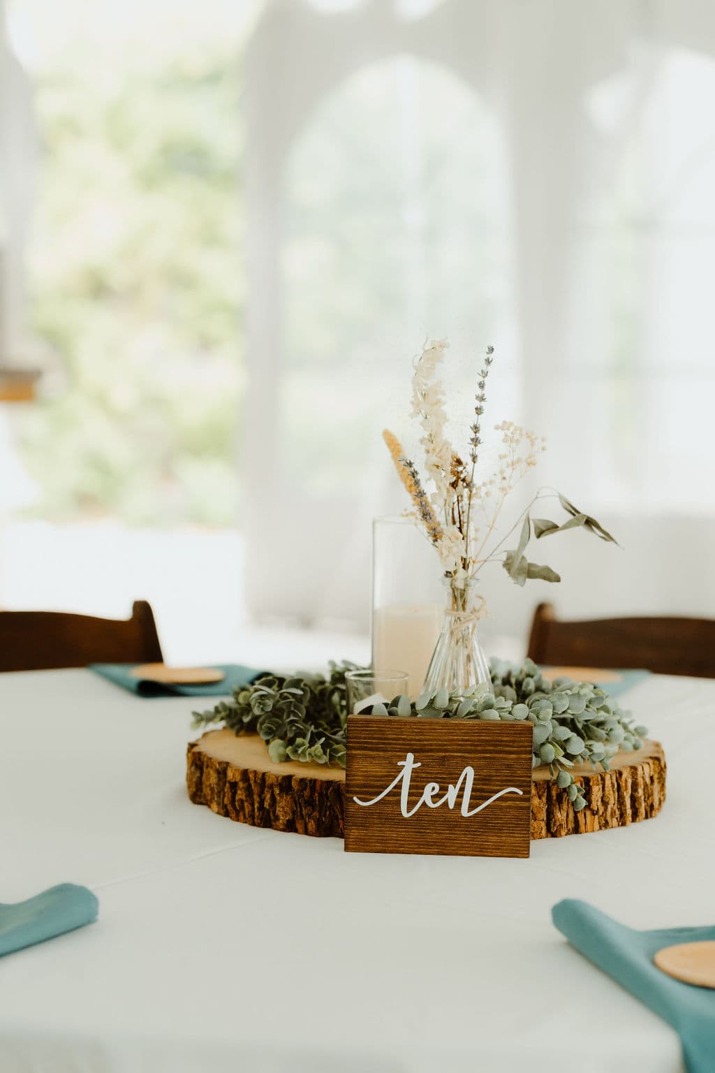 A rustic table centerpiece featuring dried flowers in a glass vase, surrounded by greenery and a wooden "ten" sign.