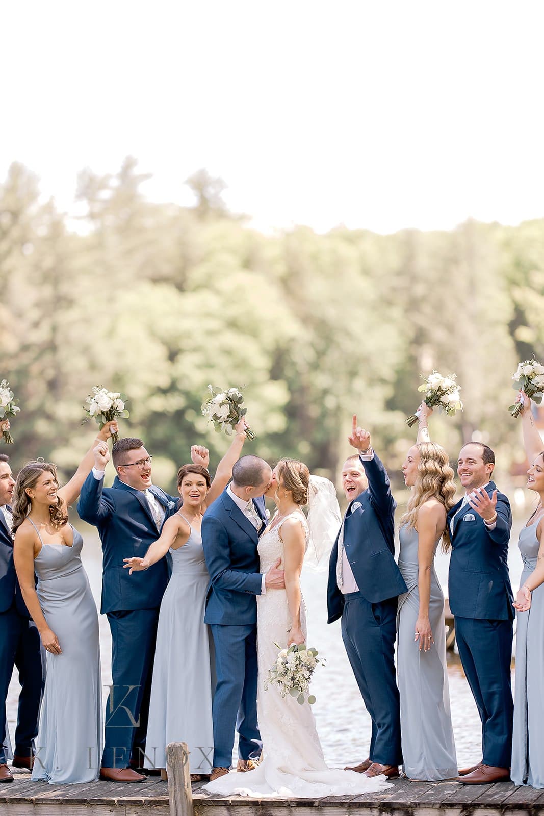 A bride and groom kiss while surrounded by their joyful wedding party on a lakeside dock.