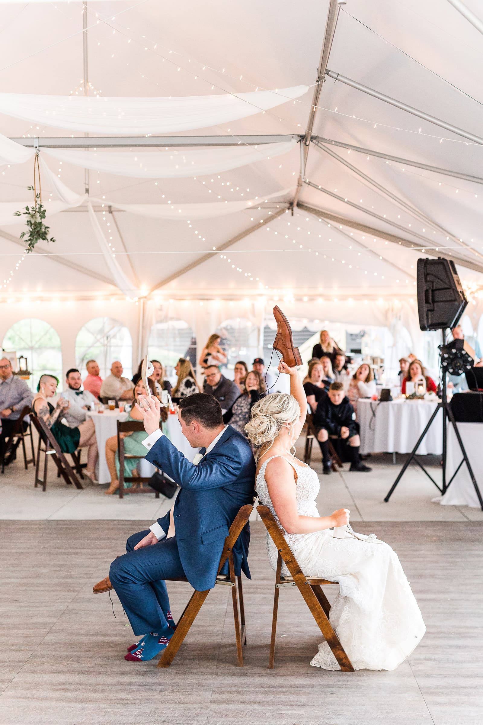 A bride and groom play a game sitting back-to-back in a festive tent filled with guests.