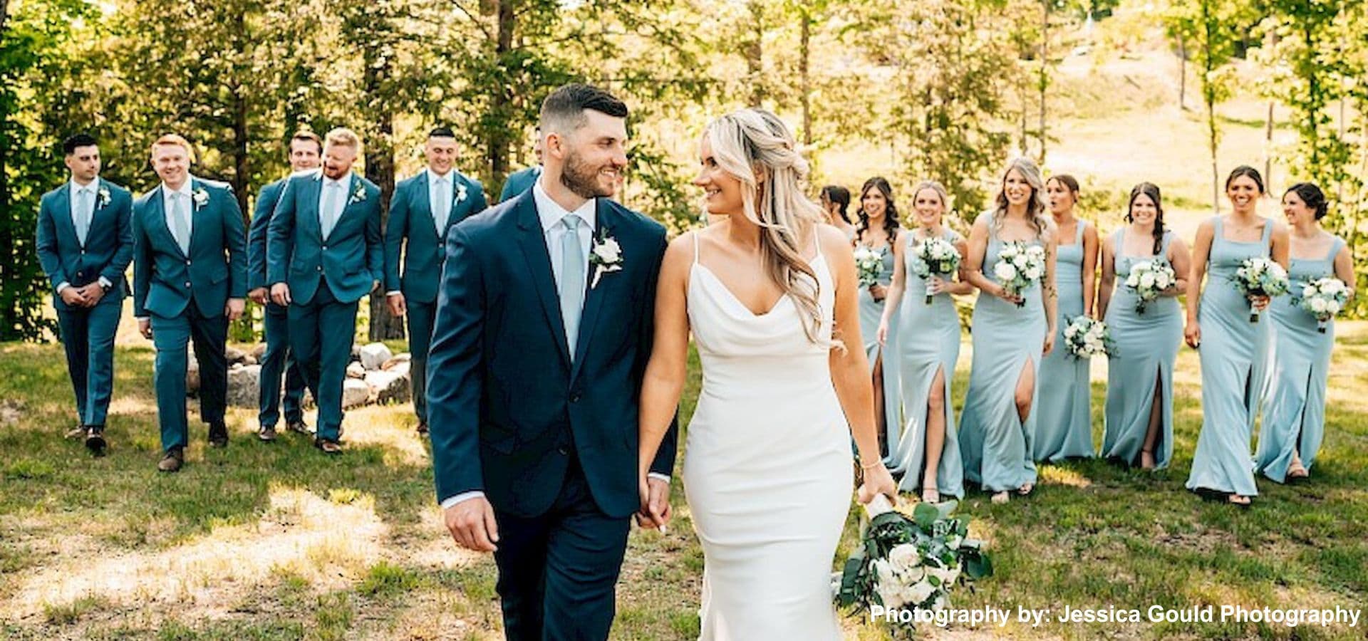 A bride and groom walk hand-in-hand, surrounded by their wedding party in elegant attire, amidst a lush green setting.