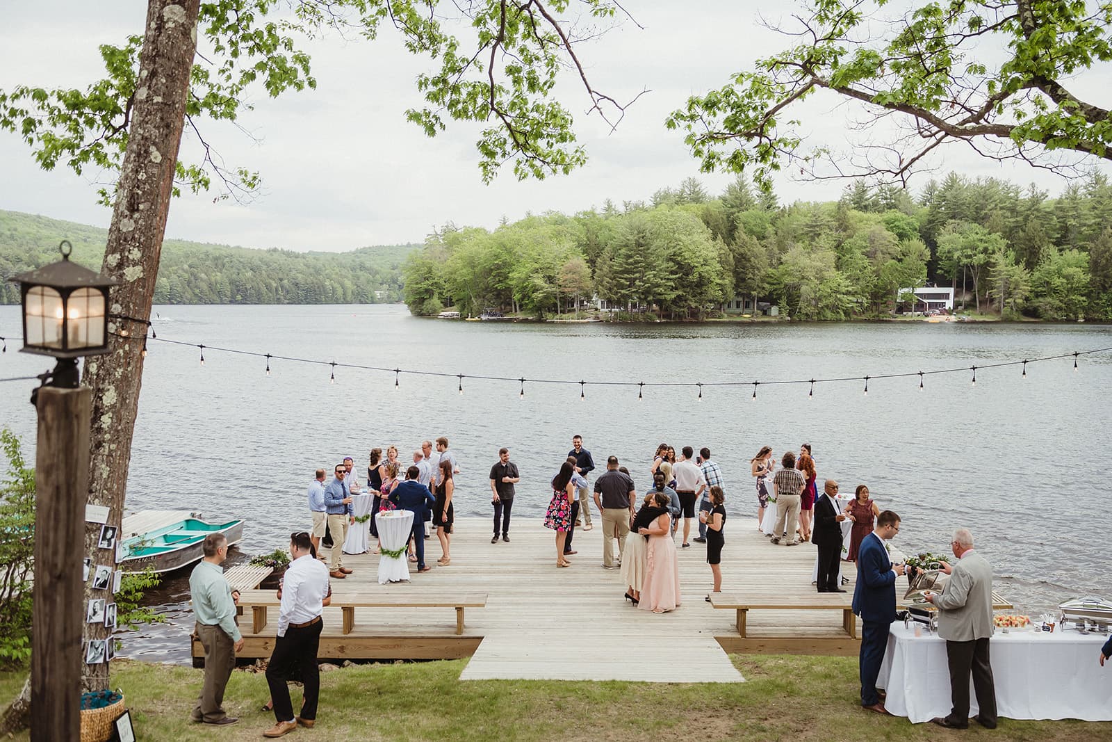 A lakeside gathering with guests mingling and dining on a wooden dock surrounded by greenery.